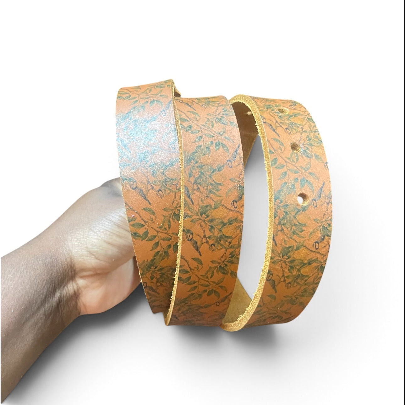 A hand holding a coiled tan leather belt with a green botanical pattern, shown against a plain white background.