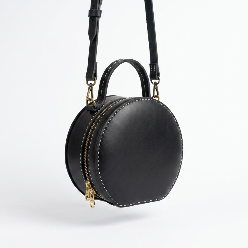 A black leather round crossbody bag with a top handle, gold-tone hardware, and white contrast stitching, shown against a clean white background.