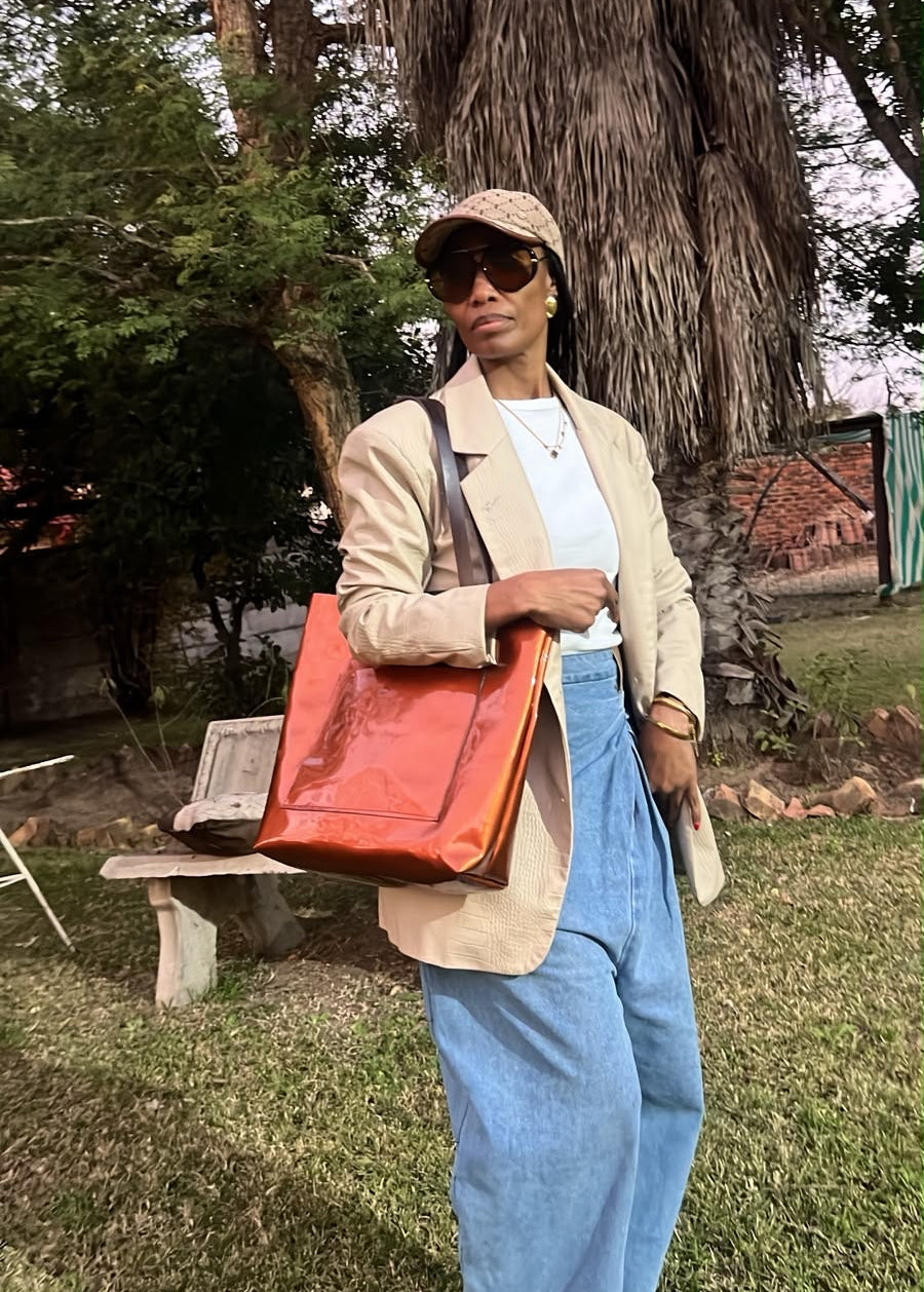 A person standing outdoors in front of a large tree, wearing a beige blazer, light blue jeans, a beige cap, and sunglasses while carrying a large orange-red tote bag.