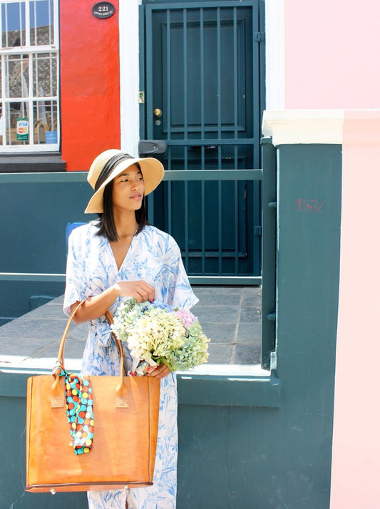 A woman standing on a doorstep carrying a large brown tote bag with multicolored flowers in it, and wearing a blue dress and a wide-brimmed hat.