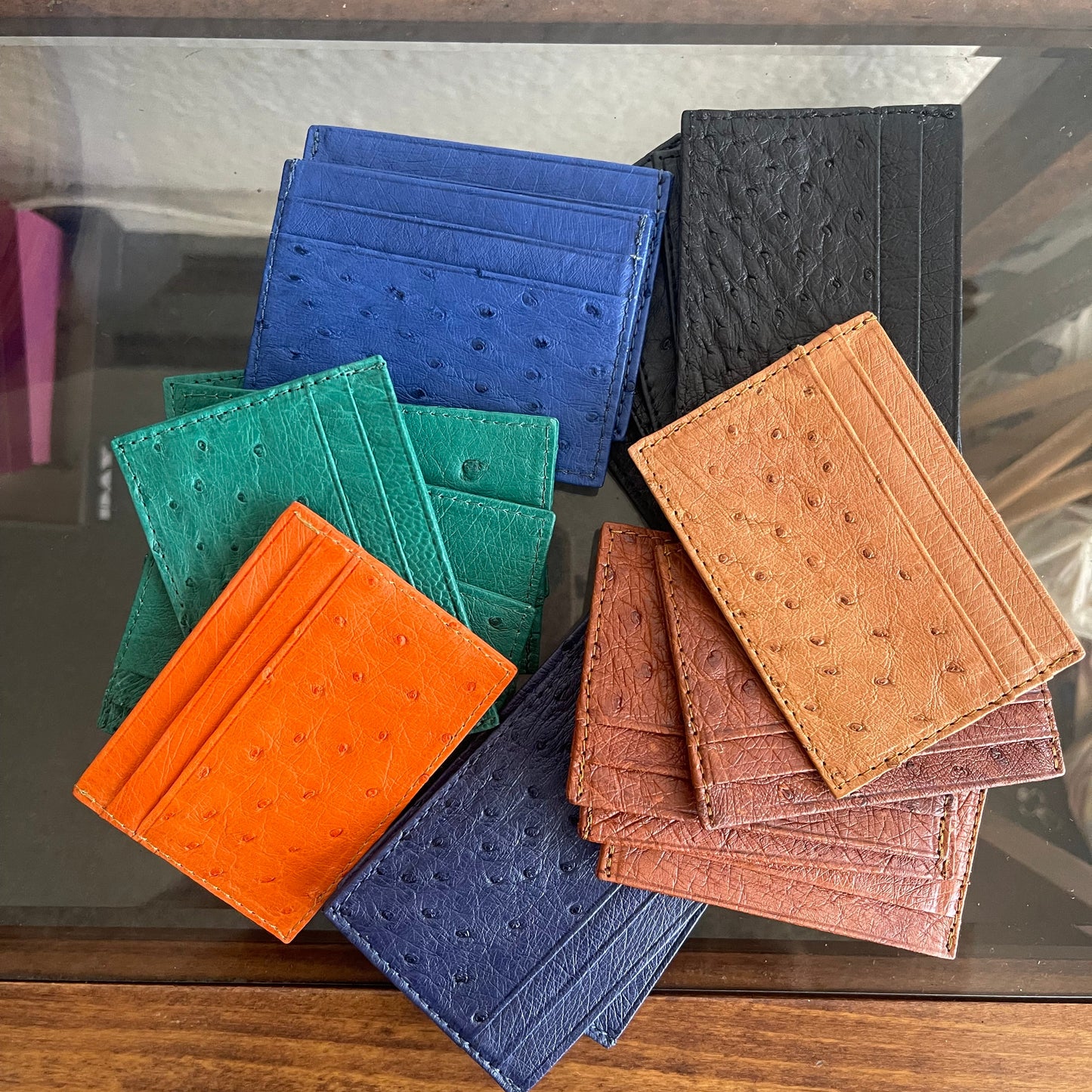A group of textured leather card holders and wallet-like accessories in multiple colors, including black, tan, reddish-brown, blue, teal, and orange, arranged on a glass surface with a wooden frame visible around it.