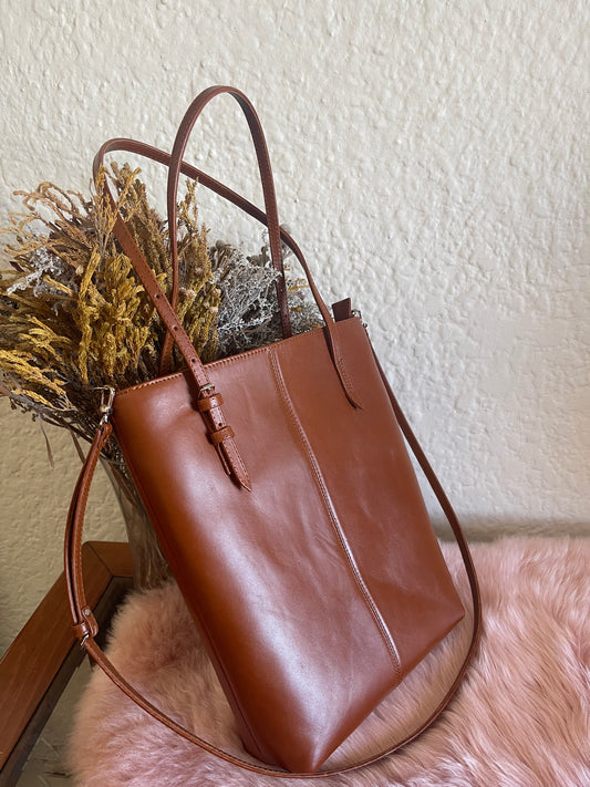 A person standing by a window, holding a brown leather tote bag.